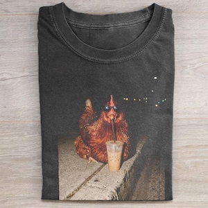 May include: A charcoal gray crew neck t-shirt featuring a graphic print of a brown chicken wearing black sunglasses and drinking from a clear plastic cup with a straw. The chicken is positioned on a concrete ledge against a dark background.