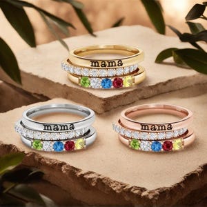 Può includere: Tre anelli impilati in oro, argento e oro rosa. Ogni anello presenta la parola "mama" e una fascia di pietre trasparenti. Gli anelli inferiori hanno una fila di pietre preziose colorate: verde, blu, rosso e giallo.