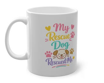 May include: A white ceramic mug featuring a colorful graphic with the text My Rescue Dog Rescued Me. The design includes a cartoon dog face, pink and brown paw prints, and small yellow and pink hearts arranged in a vertical, multi-colored layout.