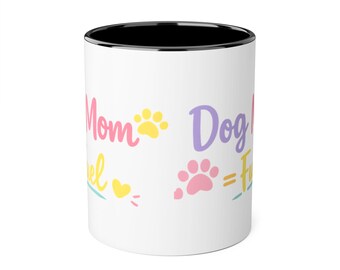 May include: A white ceramic mug with a black interior rim. The exterior features colorful text reading Dog Mom and Mom, accompanied by yellow and pink paw print graphics, a yellow heart, and an equals sign, creating a playful, multi-colored design.