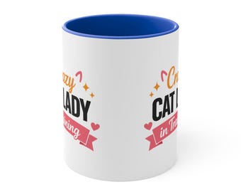 May include: White ceramic mug with a blue interior and the words "Crazy Cat Lady in Training" printed in black and pink. The mug has a pink banner with hearts.