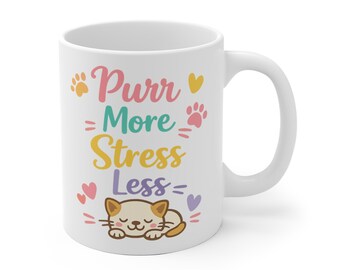 May include: A white ceramic mug featuring the colorful text Purr More Stress Less, decorated with small paw prints, hearts, and a graphic illustration of a sleeping cat with brown ears and a tan body, positioned at the bottom of the design.
