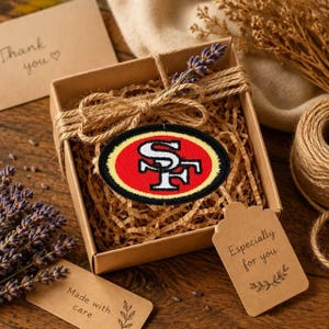 Puede incluir: Un parche bordado de forma ovalada con el logotipo rojo, dorado y blanco de los San Francisco 49ers. El parche está dentro de una caja de regalo cuadrada de cartón marrón rellena de papel picado, atada con cuerda de yute y lavanda seca.