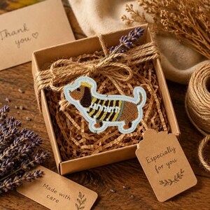 Puede incluir: Un parche bordado de un perro salchicha marrón y beige con un suéter de rayas amarillas y negras, colocado dentro de una caja de regalo cuadrada de cartón con papel picado, atada con cordel y decorada con una rama de lavanda seca.
