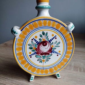 Puede incluir: Una cantimplora redonda de cerámica con un diseño floral. La cantimplora tiene una base blanca con un motivo floral central en rojo, verde y azul. Tiene un borde amarillo y azul y pequeños pies. La cantimplora tiene unos 18 cm de diámetro.