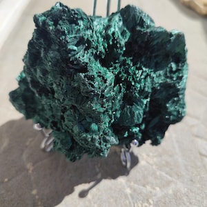 Peut inclure: Un spécimen minéral de malachite vert foncé brut avec une surface texturée et cristalline. La pierre est présentée sur un support en fil métallique argenté doté de trois petits pieds arrondis et d'un bras de support vertical pour la maintenir.