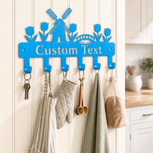 Può includere: Un appendiabiti da parete in metallo blu brillante con un design decorativo ritagliato raffigurante un mulino a vento, tulipani e un ponte. La barra orizzontale mostra il testo Custom Text e include sei ganci che sostengono vari utensili.