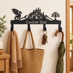 Könnte beinhalten: Eine schwarze Wandgarderobe aus Metall mit der Silhouette eines Drachen, einer Burg und eines Baumes. Die horizontale Leiste zeigt den Text Custom Text und hat vier Haken, an denen ein beiger Schal, eine geflochtene Tasche, Schlüssel und ein grüner Regenschirm hängen.