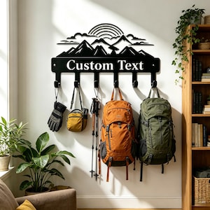 May include: A black metal wall-mounted coat rack featuring a mountain range and sun silhouette design above the text Custom Text. The rack includes six hooks holding various outdoor gear, including a glove, a small bag, trekking poles, and two backpacks.