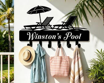 May include: A black metal wall-mounted rack featuring a silhouette of two lounge chairs under an umbrella above the text Winston's Pool. The rack includes six hooks holding a straw hat, a light blue jacket, a checkered tote bag, and a striped towel.