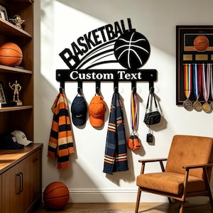 May include: A black metal wall-mounted rack featuring the word BASKETBALL, a basketball graphic, and the text Custom Text. The rack includes six hooks holding a striped scarf, two baseball caps, a patterned scarf, a lanyard, and a small bag.