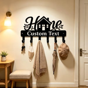 May include: A black metal wall-mounted key holder featuring a house silhouette and the word "Home" in script lettering. Below the house, a horizontal bar displays the text "Custom Text" and includes six hooks for hanging items like keys, a mask, a scarf, an umbrella, and a cap.