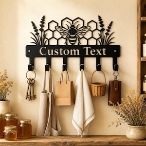 May include: A black metal wall-mounted hook rack featuring a laser-cut design of a bee, honeycomb patterns, and sprigs of lavender. The horizontal bar includes the text "Custom Text" and six downward-facing hooks for hanging various household items.