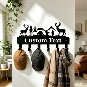 Pode incluir: Um cabide de parede em metal preto com um design de silhueta de dois veados, pinheiros e montanhas acima do texto "Custom Text". O cabide inclui seis ganchos, segurando um boné bege, um boné preto e dois cachecóis.