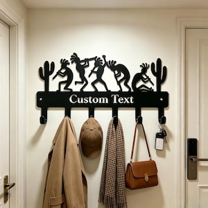 May include: A black metal wall-mounted coat rack featuring a silhouette design of five figures playing flutes and two cacti. The horizontal bar includes the text Custom Text and six hooks holding a tan coat, a baseball cap, a scarf, a bag, and keys.