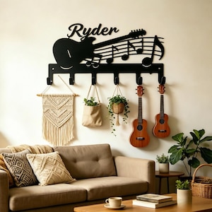 Puede incluir: Un perchero de pared de metal negro con una silueta de guitarra, notas musicales y el nombre Ryder en cursiva. El soporte tiene seis ganchos que sostienen un tapiz de macramé, una pequeña bolsa de tela con una planta y dos ukeleles de madera.