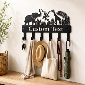 Könnte beinhalten: Eine schwarze Wandgarderobe aus Metall mit einer Dschungel-Silhouette, die einen Elefanten, einen Affen und eine Giraffe zwischen tropischen Blättern zeigt. Die horizontale Leiste trägt den Text Custom Text und hat sechs Haken für Gegenstände.
