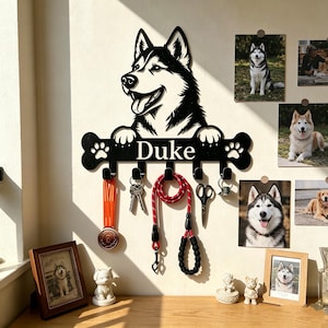 May include: A black wall-mounted metal key and leash holder featuring a cutout of a husky dog head resting its paws on a bone-shaped plaque with the name Duke printed in white, including five hooks holding a medal, keys, a red leash, and scissors.