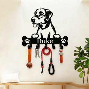 以下が含まれることがあります： 犬のイラストが描かれた白黒の壁掛け式キーホルダー。骨の形をしたプレートには「Duke」という名前が白で印字されています。プレートには5つの黒いフックがあり、メダル、鍵、赤いリード、キーホルダーが掛けられています。