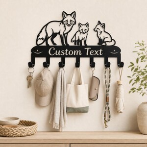 Könnte beinhalten: Eine schwarze Wandgarderobe aus Metall mit einer Silhouette eines großen Fuchses und zwei kleineren Füchsen oben. Die horizontale Leiste in der Mitte zeigt den Text Custom Text und hat sieben Haken mit Schlüsselanhänger, Kappe, Schal, Tasche, Etui, Leine und Regenschirm.