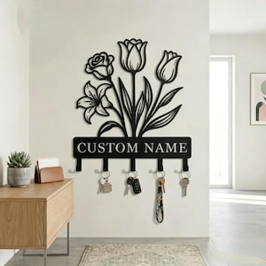 Könnte beinhalten: Ein schwarzer Wand-Schlüsselhalter aus Metall mit einem dekorativen Blumendesign aus einer Rose, Tulpen und Lilien. Die rechteckige Basis enthält den Text CUSTOM NAME und fünf Haken, an denen mehrere Schlüsselbunde hängen.