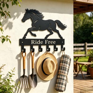 Op de afbeelding: Een zwarte metalen wandhaak met het silhouet van een galopperend paard boven de tekst "Ride Free". Het rek heeft zes naar beneden gebogen haken, waaraan een tuinvork, een schep, een strooien hoed en een geruite deken zijn opgehangen.