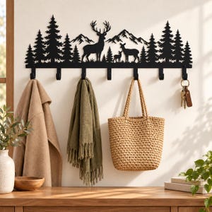May include: A black metal wall-mounted coat rack featuring a silhouette of a forest with pine trees, mountains, and a deer family. The rack includes seven hooks for hanging items, with a tan coat, olive green scarf, woven tote, and keys attached.
