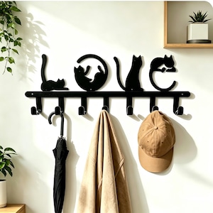 Könnte beinhalten: Eine schwarze Wandgarderobe aus Metall, die das Wort LOVE bildet, wobei die Buchstaben aus Katzensilhouetten bestehen. Die Garderobe hat sieben nach unten gebogene Haken. Ein schwarzer Regenschirm, eine beige Jacke und eine beige Kappe hängen daran.