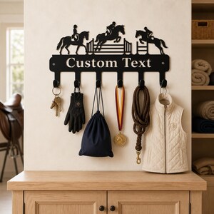 Könnte beinhalten: Eine schwarze Wandgarderobe aus Metall mit einer Silhouette von drei Pferden und Reitern, die über Hindernisse springen. Die horizontale Leiste zeigt den Text Custom Text und hat sechs Haken mit Schlüsseln, Handschuhen, einer Tasche, einer Medaille und einer Weste.