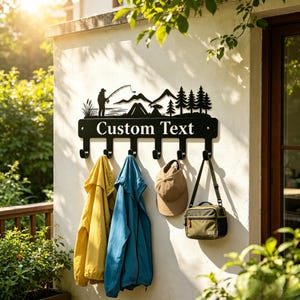 May include: A black metal wall-mounted coat rack featuring a silhouette of a person fishing near mountains and pine trees. The horizontal bar displays the text Custom Text and includes six hooks holding a yellow jacket, blue jacket, tan cap, and bag.