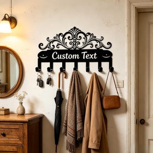 Könnte beinhalten: Eine schwarze Wandgarderobe aus Metall mit Schlüsselhaken, verziert mit einem ornamentalen Schnörkeldesign und dem weißen Schriftzug "Custom Text". Die Garderobe verfügt über sieben Haken, an denen Schlüssel, ein Regenschirm, ein Schal und ein Mantel hängen.