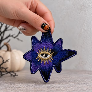 May include: A star-shaped embroidered patch featuring a gradient of deep blue and purple threads. The center displays an eye motif stitched in gold thread with a small white bead at the pupil, surrounded by a border of tiny dark blue beads.