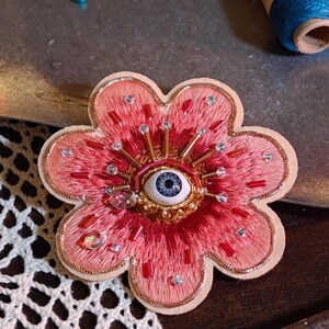 Könnte beinhalten: Handgefertigte, blütenförmige Brosche mit zentralem Augendesign. Die Brosche zeigt eine rosa bestickte Blume mit goldenen Akzenten, roten Perlen und klaren Kristallen. Das Auge hat eine blaue Iris und ist von goldenen Strahlen umgeben.