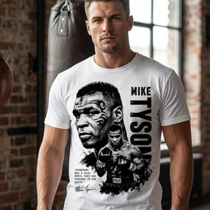 May include: White t-shirt featuring a black and white graphic of Mike Tyson. The design includes Tyson's portrait, a boxing pose, and the text "MIKE TYSON". The shirt also has a quote and signature. A watch is visible on the wrist.