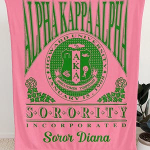 Op de afbeelding: Een roze fleece deken met groene tekst en afbeeldingen. Het ontwerp bevat de woorden Alpha Kappa Alpha, Howard University, Sorority Incorporated en de naam Soror Diana, naast een centraal embleem met een schild, rozenmotieven en jaartallen.