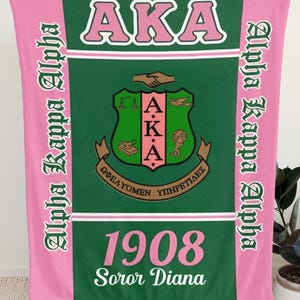 Op de afbeelding: Een rechthoekige fleece deken met een donkergroen midden en roze zijpanelen. De tekst AKA, 1908 en Soror Diana is gedrukt in roze en wit. Het midden toont een wapenschild met symbolen en aan de zijkanten staat de tekst Alpha Kappa Alpha.