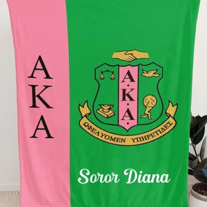 Op de afbeelding: Een rechthoekige fleece deken met een verticale roze streep aan de linkerkant met de zwarte letters AKA verticaal gestapeld. Het grotere groene gedeelte toont een centraal wapenschild met de letters AKA, een handdruk en Griekse tekst, met de naam Soror Diana in wit schrift eronder.