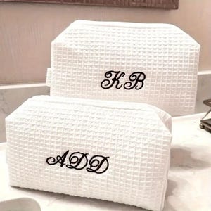 Peut inclure: Deux trousses de toilette rectangulaires blanches en tissu gaufré texturé. Le plus grand sac est brodé des lettres script noires KB, tandis que le plus petit sac à l'avant est brodé des lettres script noires ADD.