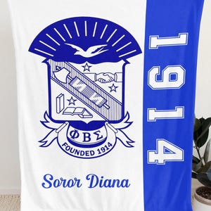 Op de afbeelding: Een witte en koningsblauwe fleece deken met een groot wapenschild met symbolen van een vogel, een handdruk en een boek. De tekst luidt Phi Beta Sigma, Founded 1914 en Soror Diana. Een verticale blauwe baan aan de rechterkant toont het jaartal 1914.