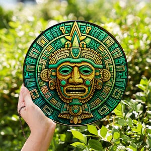 Op de afbeelding: Een ronde wanddecoratie met een gedetailleerd gezicht met een hoofdtooi, in tinten groen, geel en goud. Het ontwerp bevat ingewikkelde geometrische patronen. Het kunstwerk wordt tegen een achtergrond van groen gebladerte gehouden.