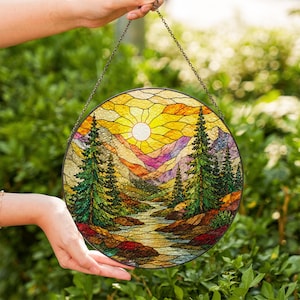 Könnte beinhalten: Ein runder Buntglas-Sonnenfänger mit einer Landschaftsszene, die eine leuchtend gelbe Sonne, violette und orangefarbene Berge, einen Fluss und grüne Tannen zeigt. Das Stück ist in schwarzes Metall gefasst und hängt an einer dünnen Metallkette.