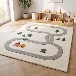 May include: A rectangular area rug in cream features a gray winding road pattern with small appliqués of green triangular trees, orange and yellow houses, and two small toy cars in blue and orange placed along the track.