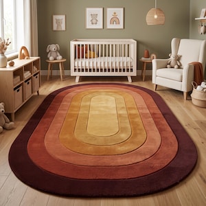 May include: An oval-shaped area rug featuring a concentric rainbow pattern in shades of yellow, orange, terracotta, and deep burgundy. The rug has a soft, plush texture and is positioned on a light-colored wooden floor in a nursery setting.