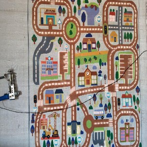May include: A rectangular rug featuring a colorful city map design with brown roads, various buildings including a hospital and school, green trees, and blue water elements. A tufting machine is attached to the left side of the fabric canvas.