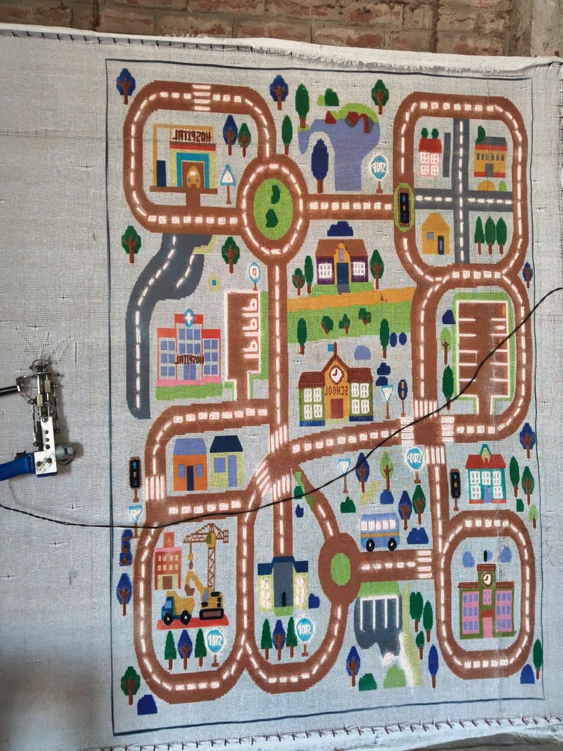 May include: A rectangular rug featuring a colorful city map design with brown roads, various buildings including a hospital and school, green trees, and blue water elements. A tufting machine is attached to the left side of the fabric canvas.