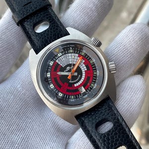 May include: A Fortis Marinemaster wristwatch with a silver-tone stainless steel case and a black textured leather strap. The dial features a black and red circular pattern, white hour markers, orange hands, and a date window at the 6 o'clock position.
