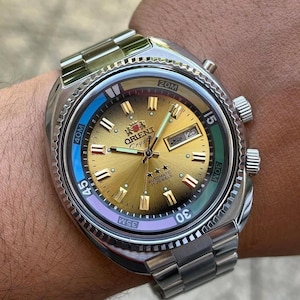 Könnte beinhalten: Eine Orient Crystal Automatikuhr mit goldfarbenem Zifferblatt im Sonnenschliff, silberfarbenen Zeigern und aufgesetzten Indizes. Die Uhr verfügt über ein Edelstahlgehäuse mit Gliederarmband, eine Datumsanzeige und eine drehbare Lünette mit blauen und violetten Akzenten.
