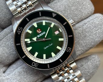 May include: A stainless steel Rado Captain Cook watch with a green dial and a black bezel. The watch features a date window at the 3 o'clock position and a stainless steel bracelet. The watch is shown on a gray surface.