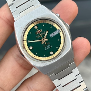 Peut inclure: Une montre-bracelet en acier inoxydable argenté avec un cadran rond vert, des index dorés et un guichet jour-date. Le cadran affiche le logo Orient, trois étoiles et le texte Crystal 21 Jewels. Elle possède un bracelet à maillons en métal argenté.