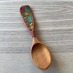 Könnte beinhalten: Handgeschnitzter Holzlöffel mit dekorativem Griff. Der Griff weist ein geometrisches Design in Teal, Gelb und Rot auf. Die Löffelschale hat eine natürliche Holzfarbe, und der Griff ist rotbraun.